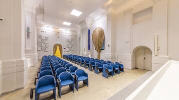 Un salón de conferencias con filas de sillas azules, paredes blancas y una lámpara moderna.