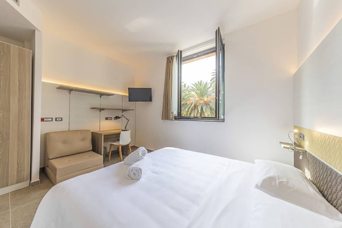 Une chambre d’hôtel équipée d’un lit, d’un bureau, d’une chaise, d’une télévision et d’une fenêtre donnant sur des palmiers.