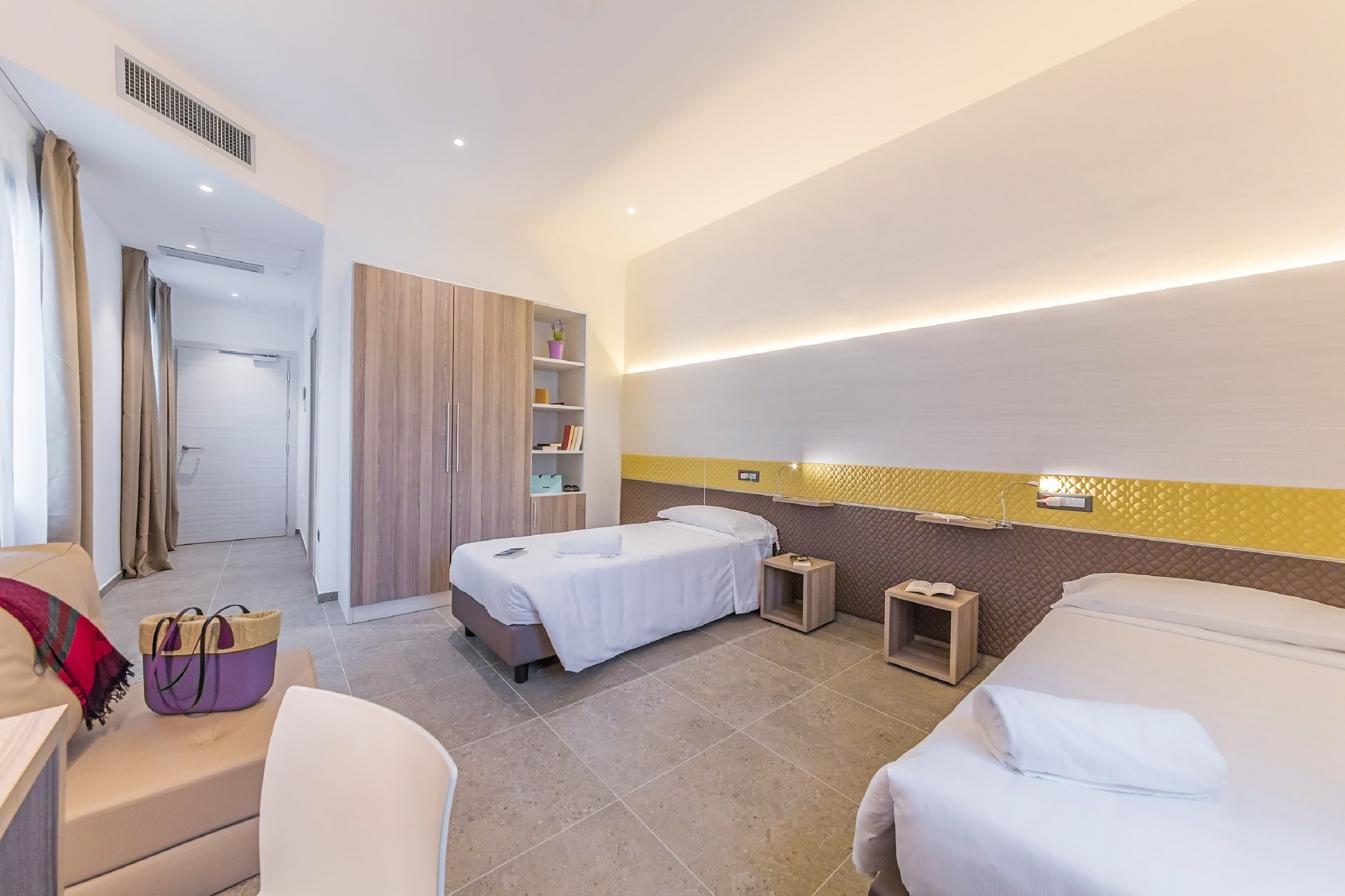 A hotel room with two beds, a desk, a chair, a wardrobe, and a small bedside table with a book.