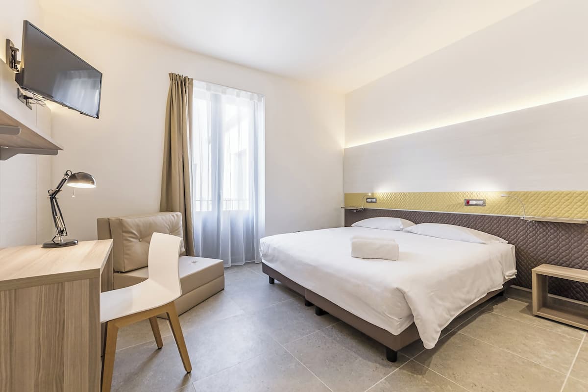 Une chambre d’hôtel moderne équipée d’un lit, d’un bureau, d’une chaise, d’un téléviseur fixé au mur et d’une fenêtre avec des rideaux.