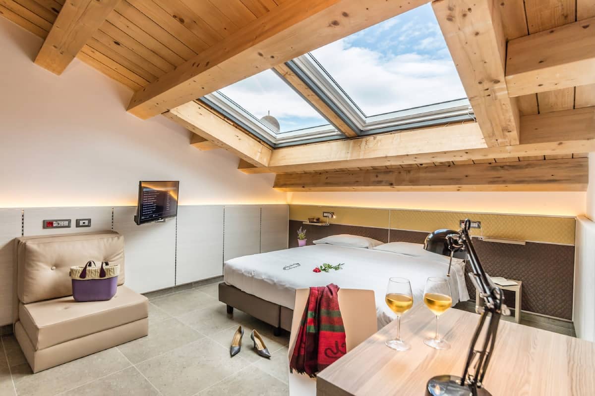 Une chambre d’hôtel avec une lucarne, un lit, un bureau avec des verres de vin et une petite table sur laquelle se trouve un sac à main.