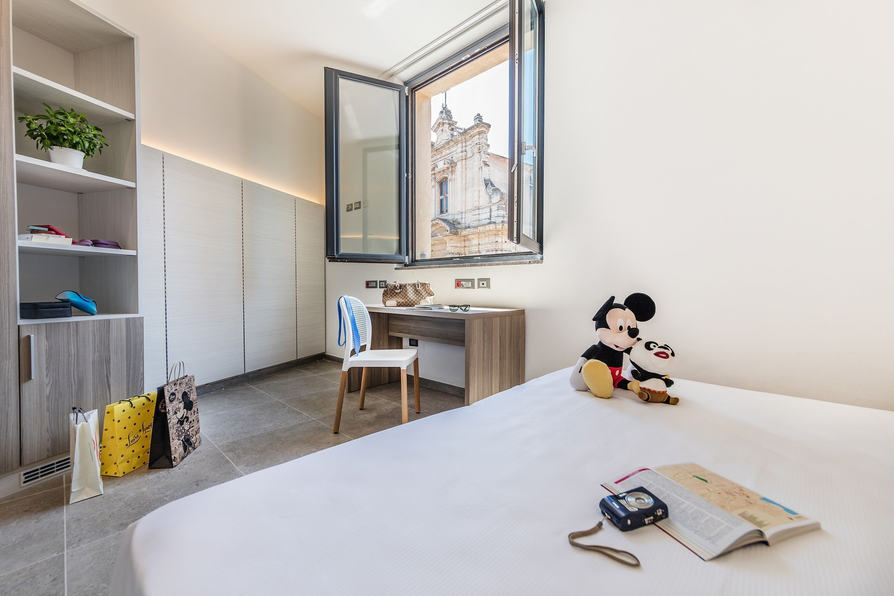 Quarto moderno com cama, escrivaninha, cadeira e janela com vista para um prédio.