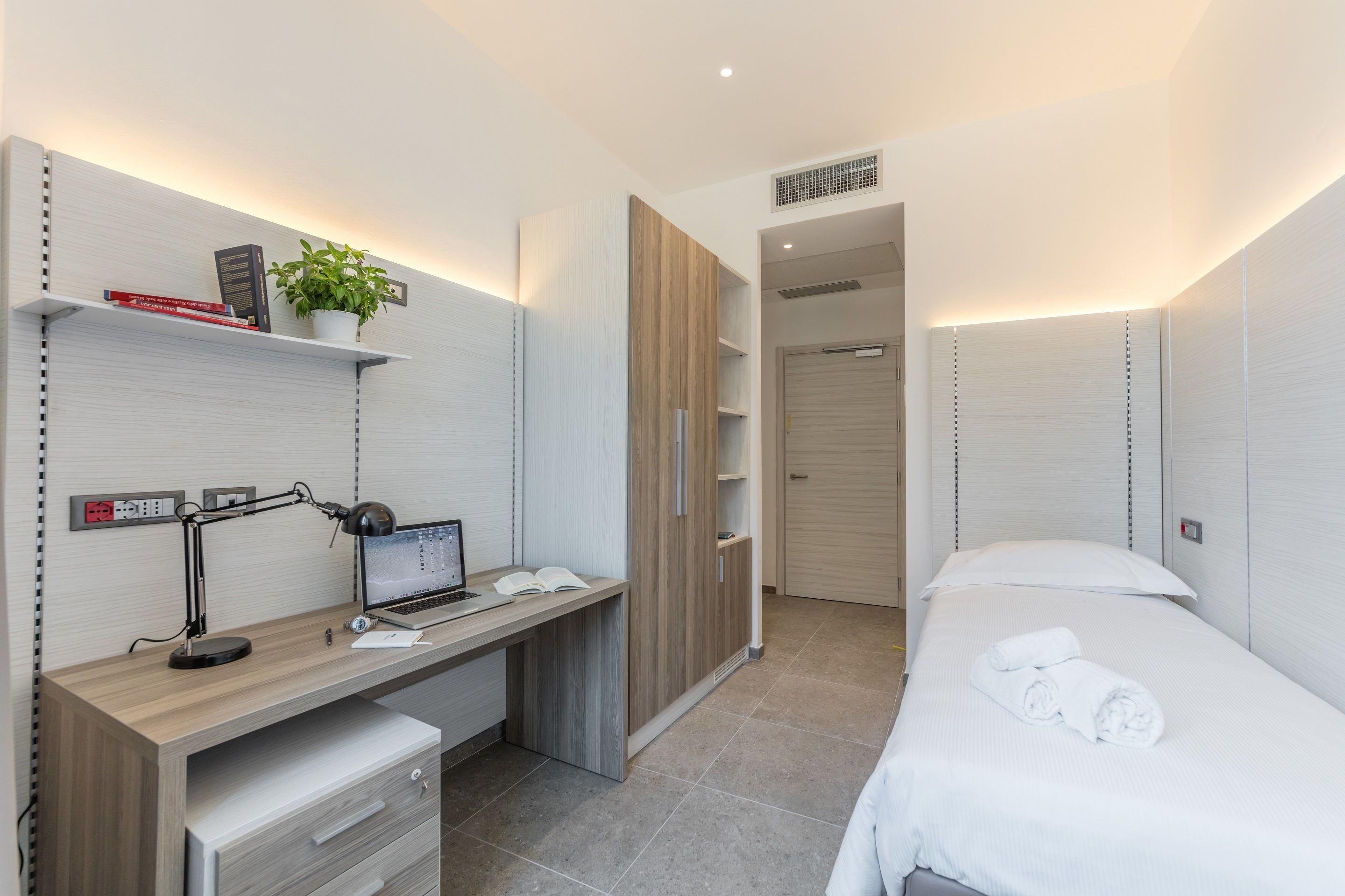 A modern bedroom with a wooden desk, a laptop, a plant, and a neatly made bed.
