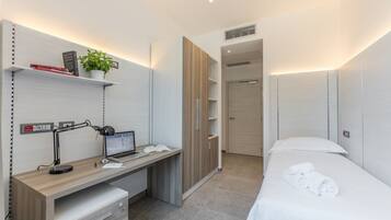 A modern bedroom with a wooden desk, a laptop, a plant, and a neatly made bed.