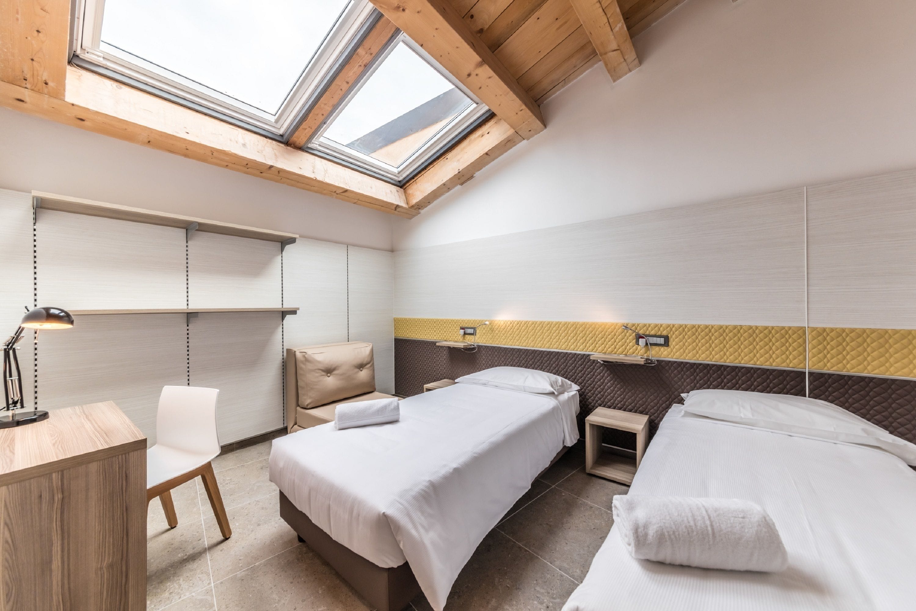 A hotel room with two beds, a desk, and a chair. There are skylights above the beds.