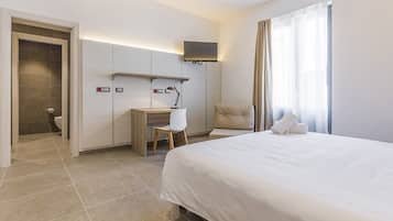 Une chambre d’hôtel comprenant un lit, un bureau avec une chaise, un téléviseur fixé au mur et une salle de bain visible à travers une porte ouverte.