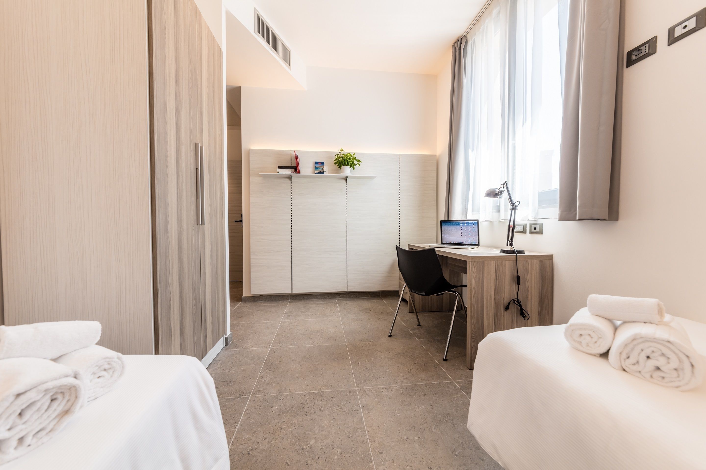 Ein Hotelzimmer mit einem Bett, einem Schreibtisch mit Computer, einem Stuhl und einem kleinen Regal mit Pflanze.