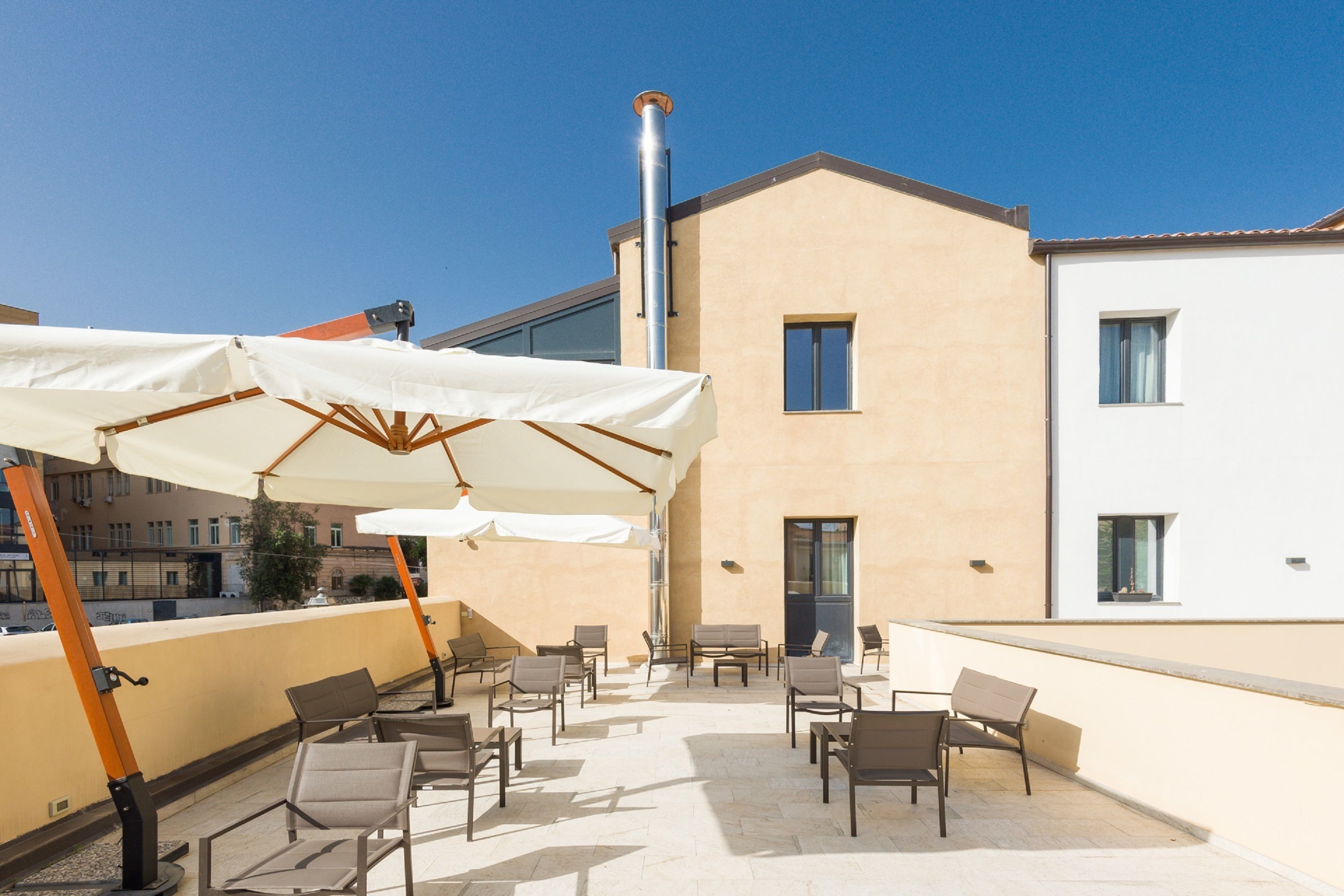 Une terrasse extérieure avec des chaises et un grand parasol, un bâtiment à la façade beige et un ciel d’un bleu limpide.