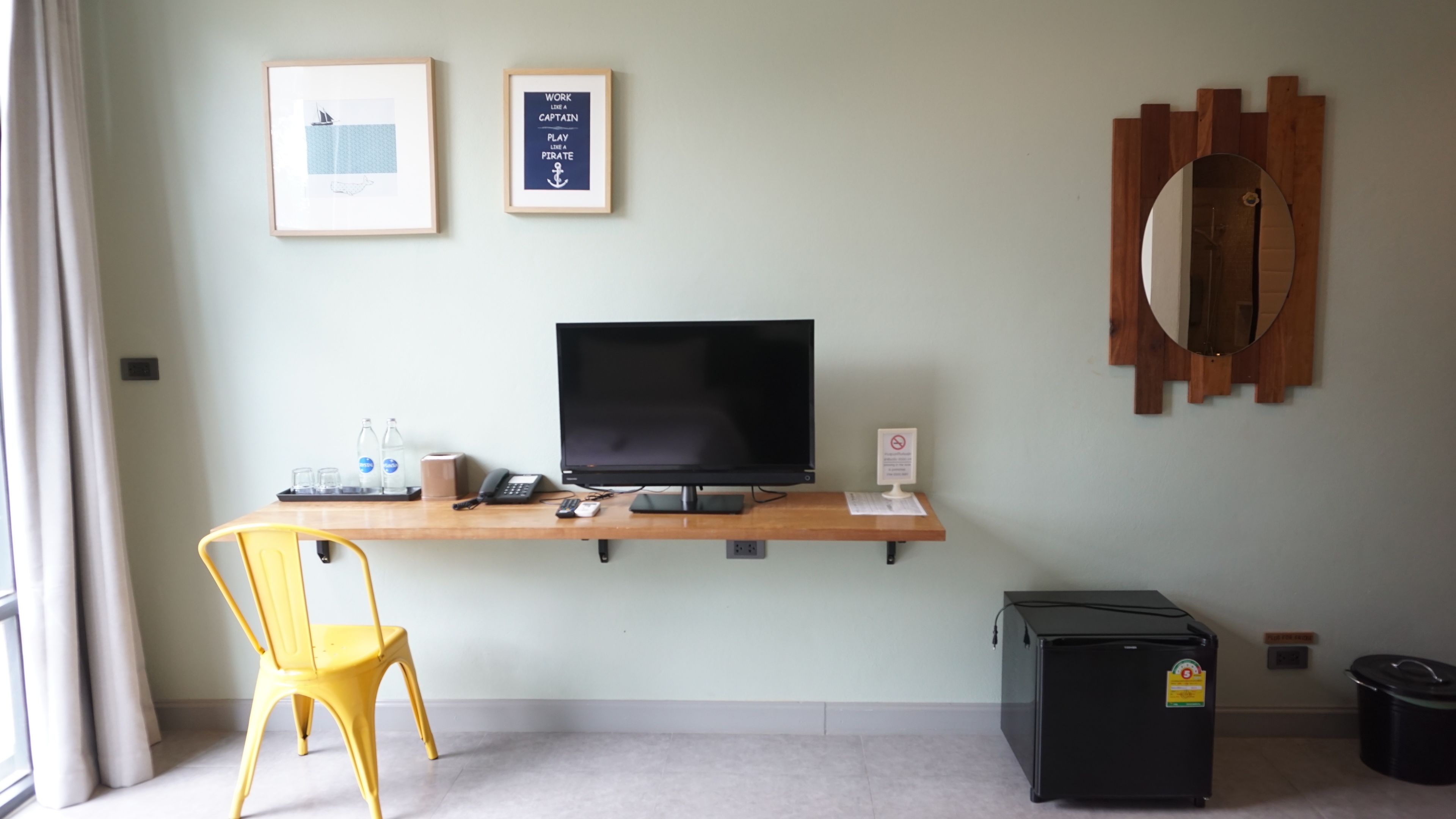 A hotel room with a desk, a television, a yellow chair, a small refrigerator, and two framed pictures on the wall.