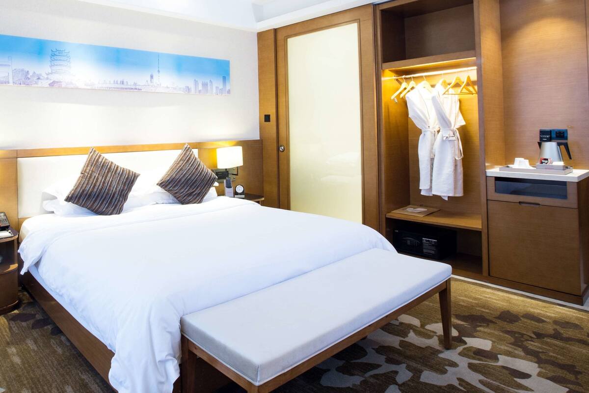 Hotellihuone, jossa on sänky, yöpöydät, naulakko kylpytakille, työpöytä kahvinkeittimellä sekä taulu seinällä.