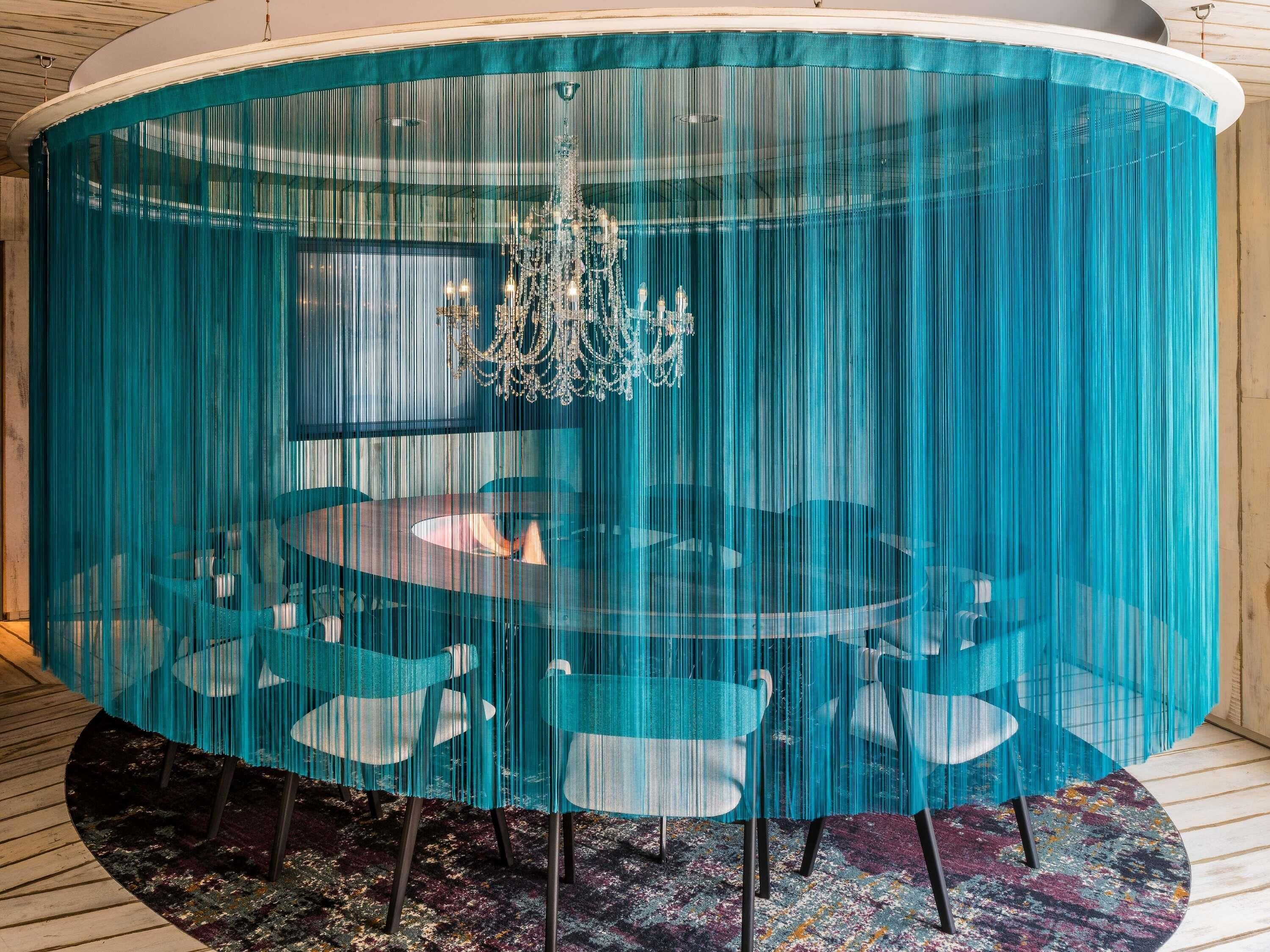 A round table with chairs, a chandelier, and teal curtains.