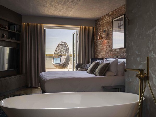 A modern bedroom with a large bed, a hanging chair, a bathtub, and a brick wall. - Novotel London Canary Wharf Hotel (London)