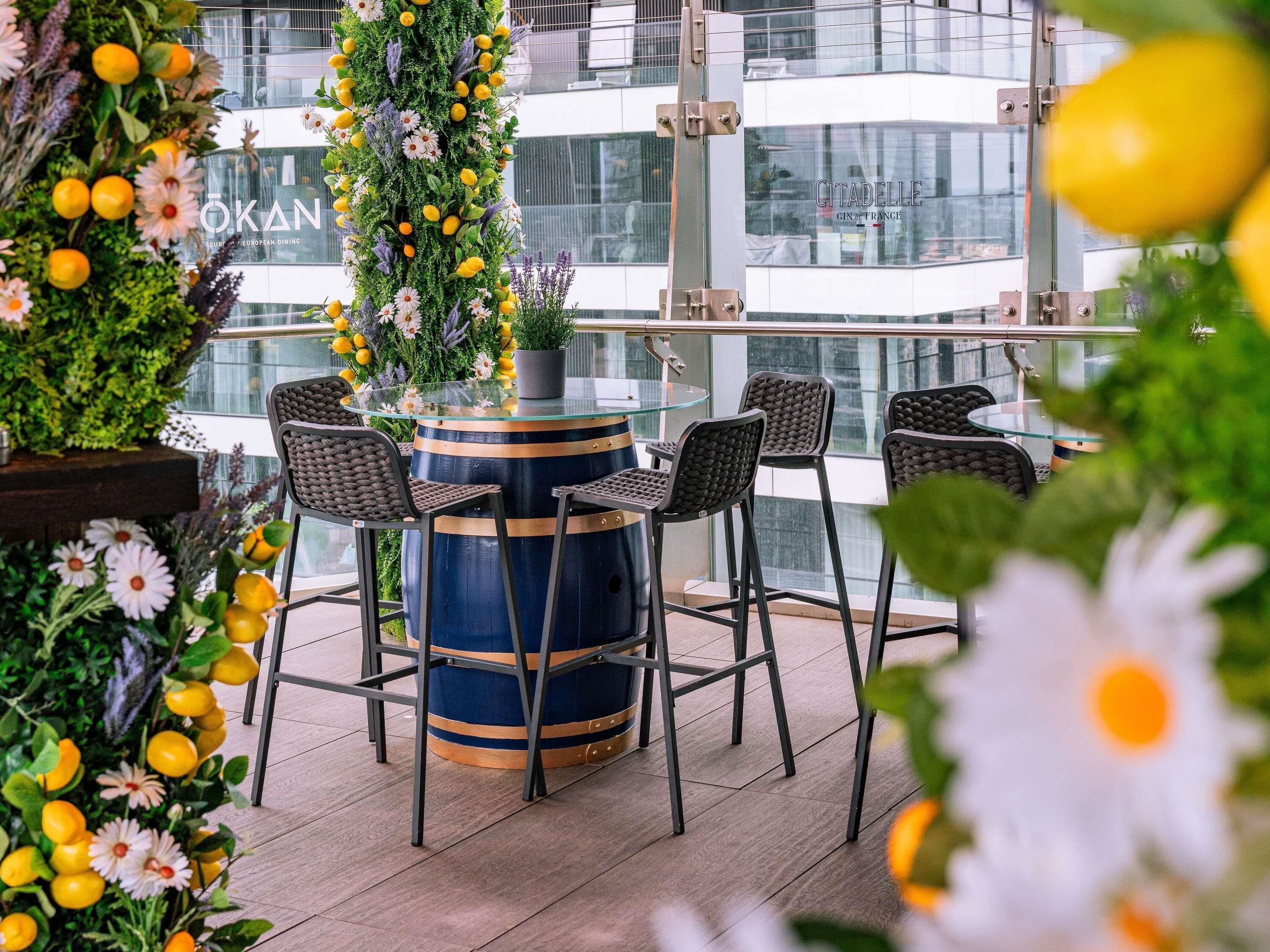 Un espace extérieur aménagé avec une table et des chaises à plateau en verre, entouré d’une végétation luxuriante et de fleurs.