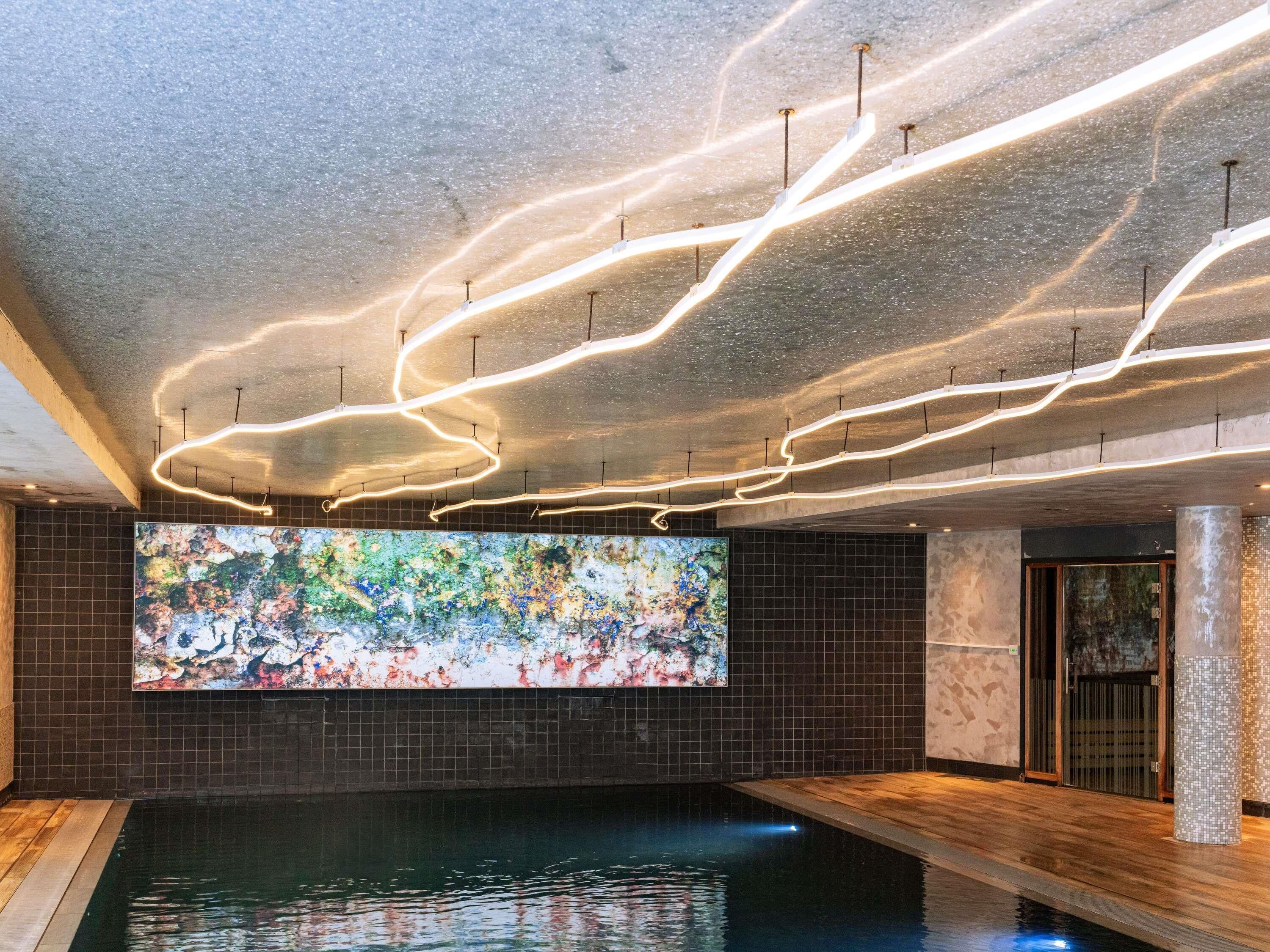 Un'area piscina moderna in interni, con un grande murale astratto, un soffitto con faretti lineari e un pavimento in legno.