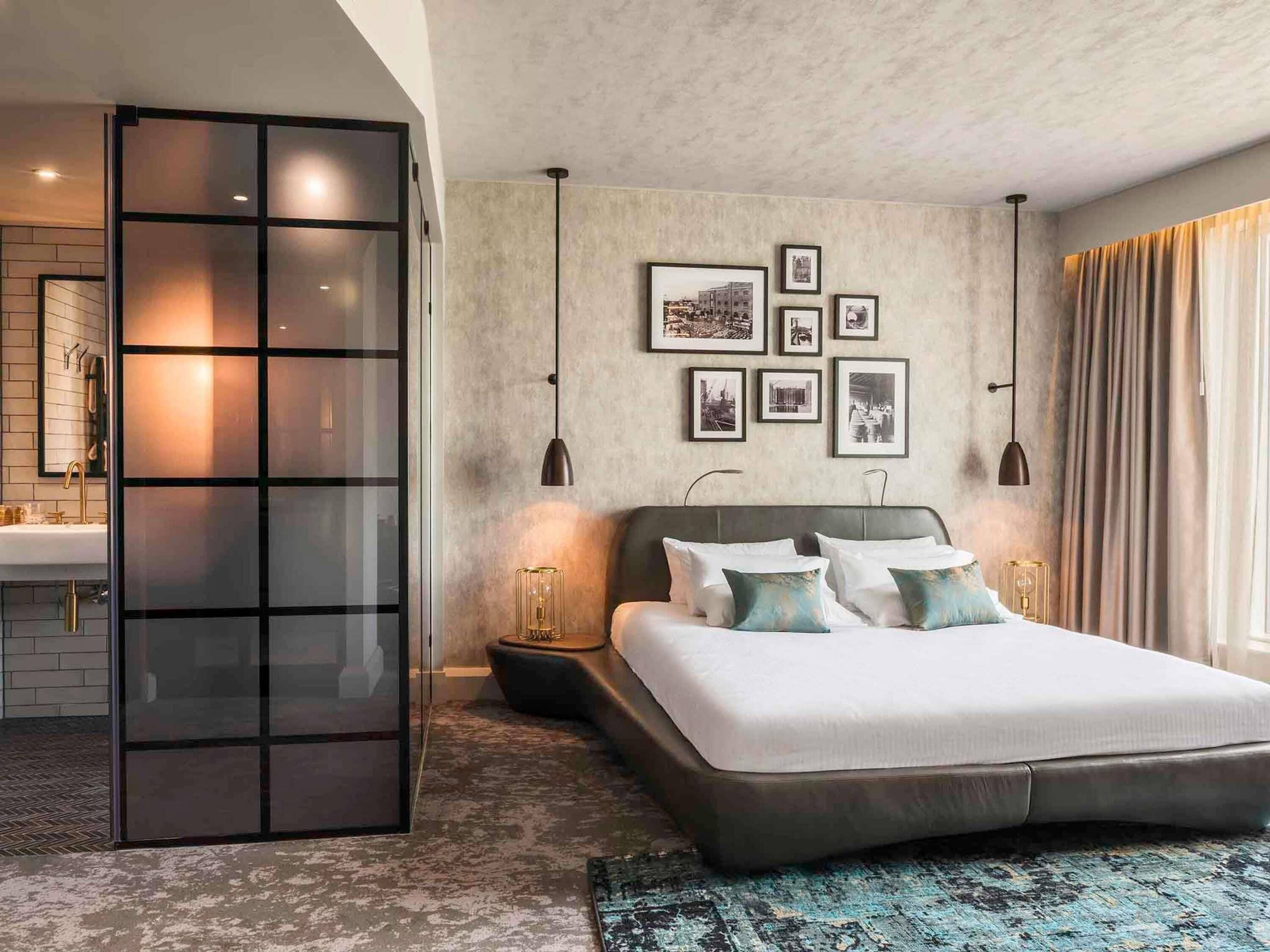 A modern bedroom with a large bed, a glass partition, a bathroom with a sink, and framed pictures on the wall.