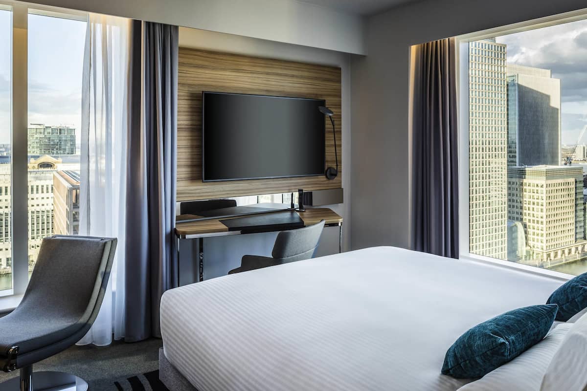 Une chambre d’hôtel moderne dotée d’un grand lit, d’un bureau avec un téléviseur à écran plat, d’une chaise et offrant une vue sur le paysage urbain grâce à de grandes fenêtres.