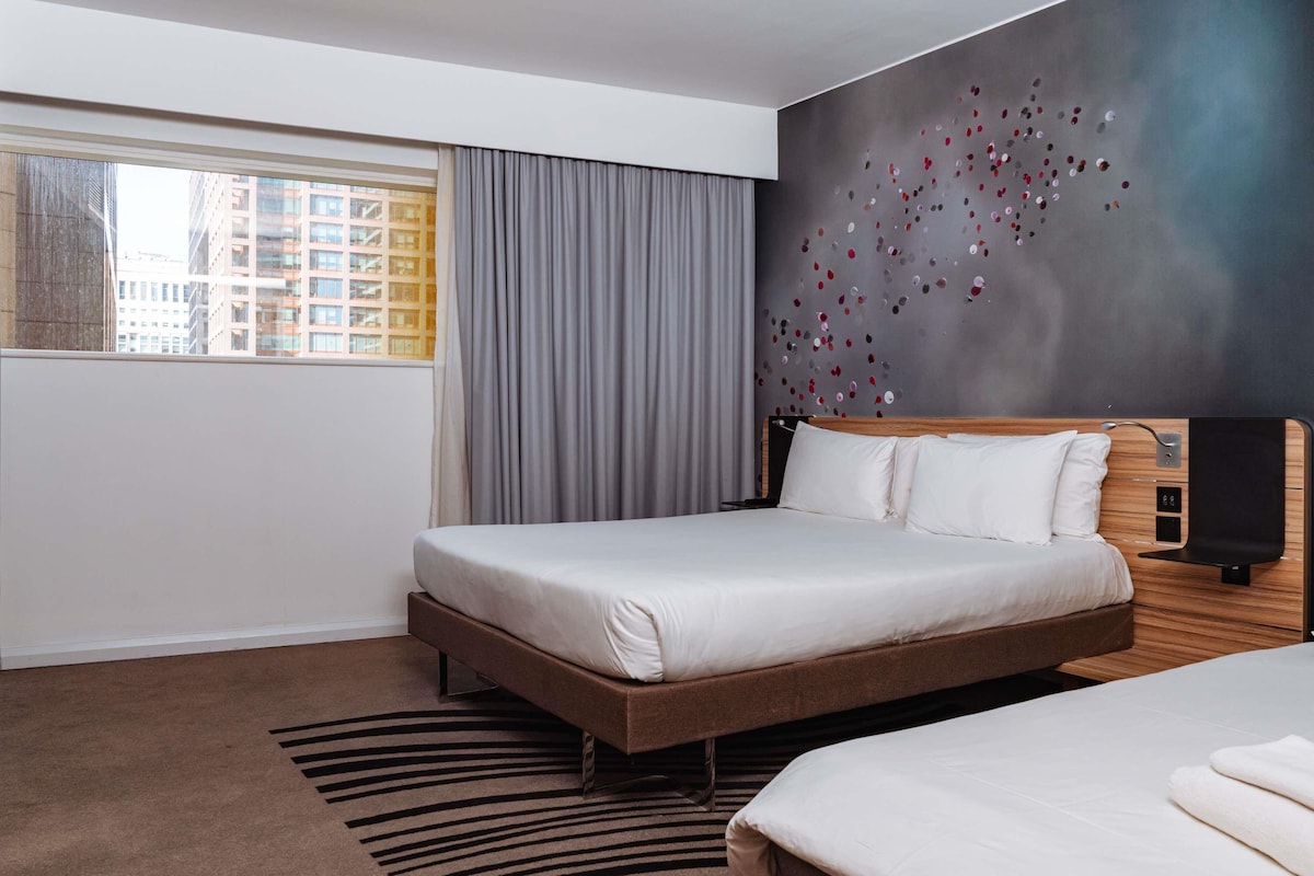 Ein Hotelzimmer mit einem großen Bett, einem kleineren Bett, einem Fenster mit Vorhängen, einer Wand mit dekorativem Muster und einem Teppich mit geometrischem Design.
