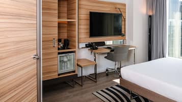 A hotel room with a bed, a desk with a chair, a television, and a minibar.