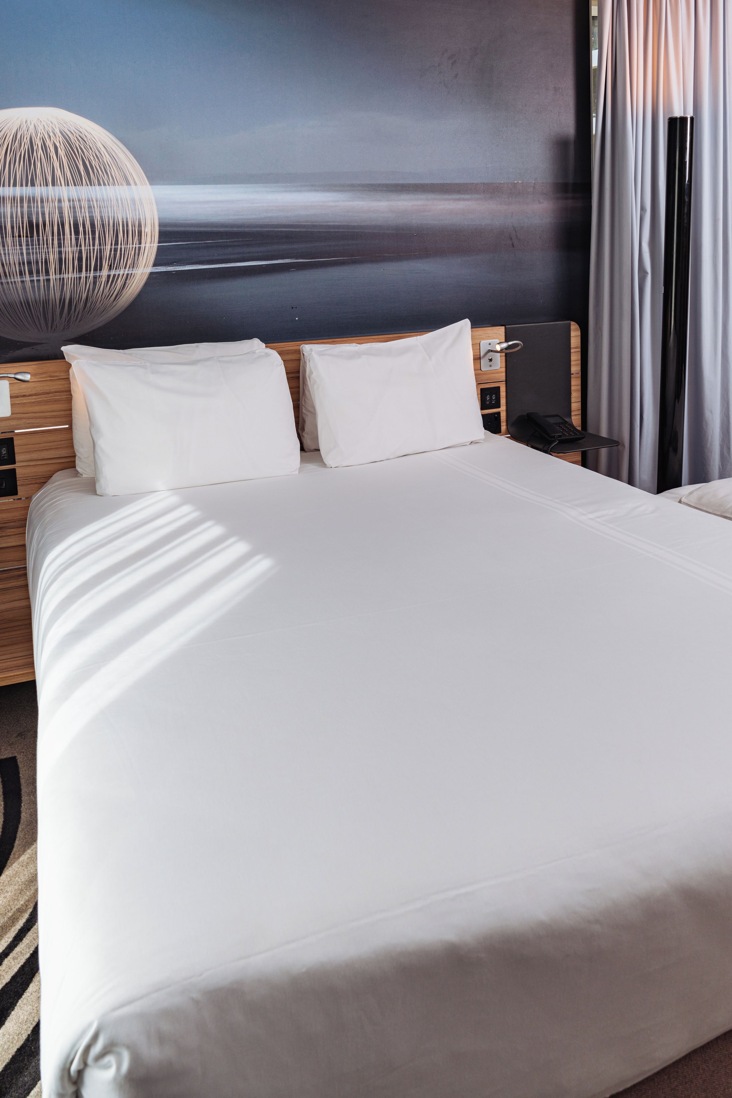 A hotel room with a large bed, a wooden headboard, a wall-mounted lamp, and a view of the sea through the window.