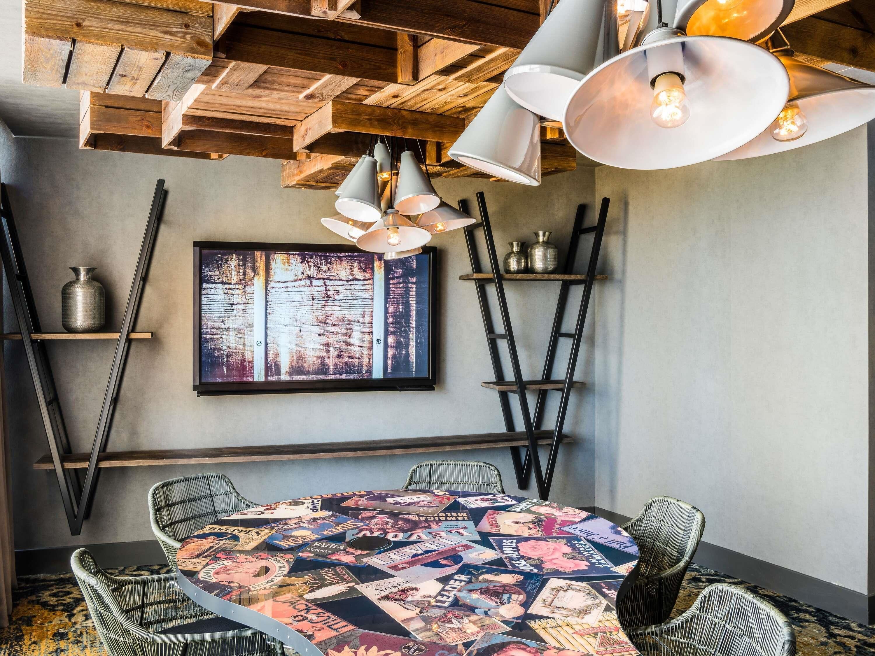 A dining area with a round table covered in magazines, metal chairs, a wooden ceiling, and pendant lights.