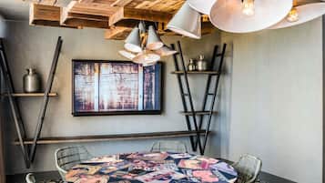 A dining area with a round table covered in magazines, metal chairs, a wooden ceiling, and pendant lights.