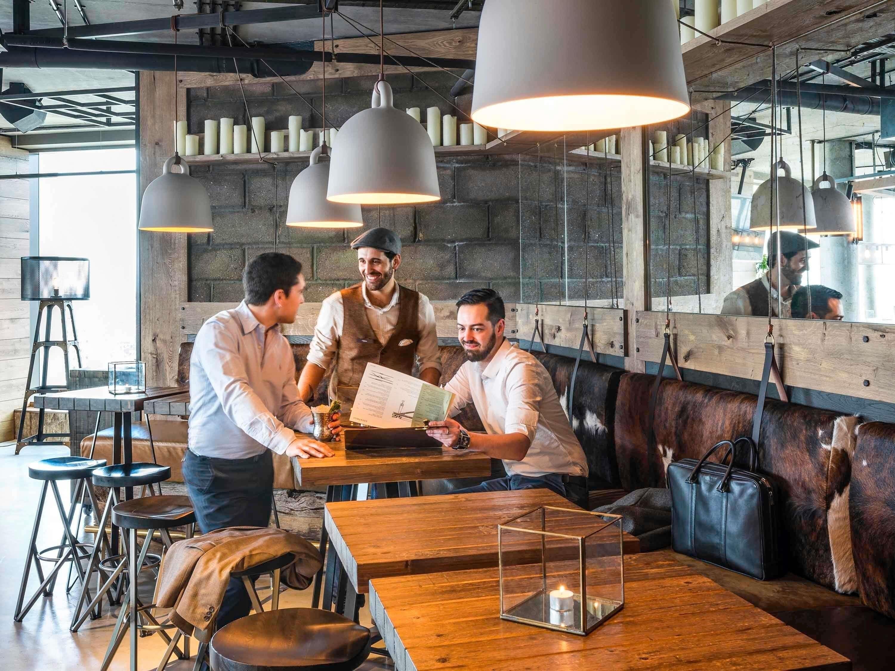 A restaurant interior with wooden tables, bar stools, hanging pendant lights, and a seating area with leather cushions.