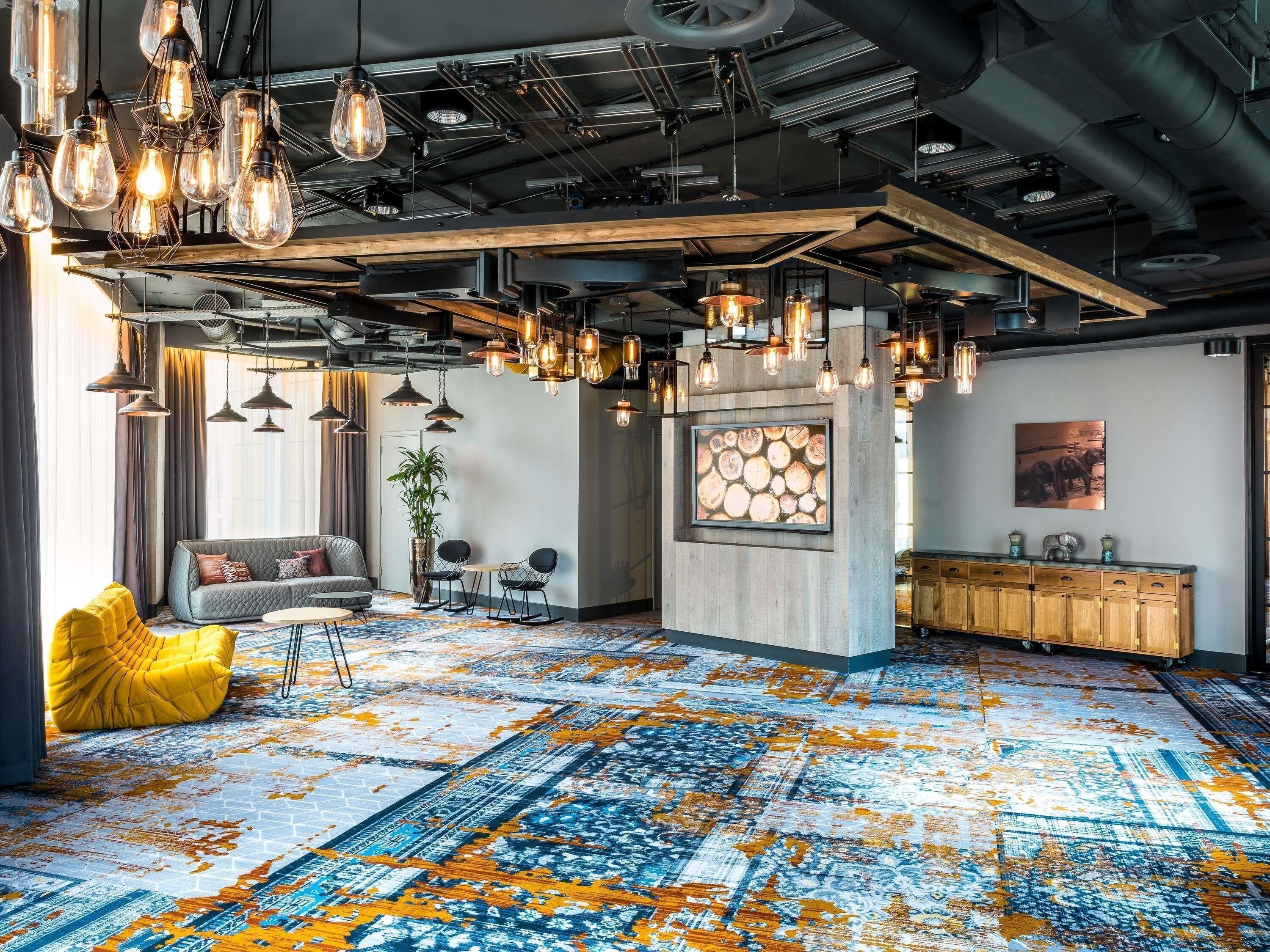 Ein modernes Interieur mit freiliegenden Decken, hängenden Leuchten im Industriedesign, einem gemusterten Teppich und einer Sitzecke mit gelbem Pouf.
