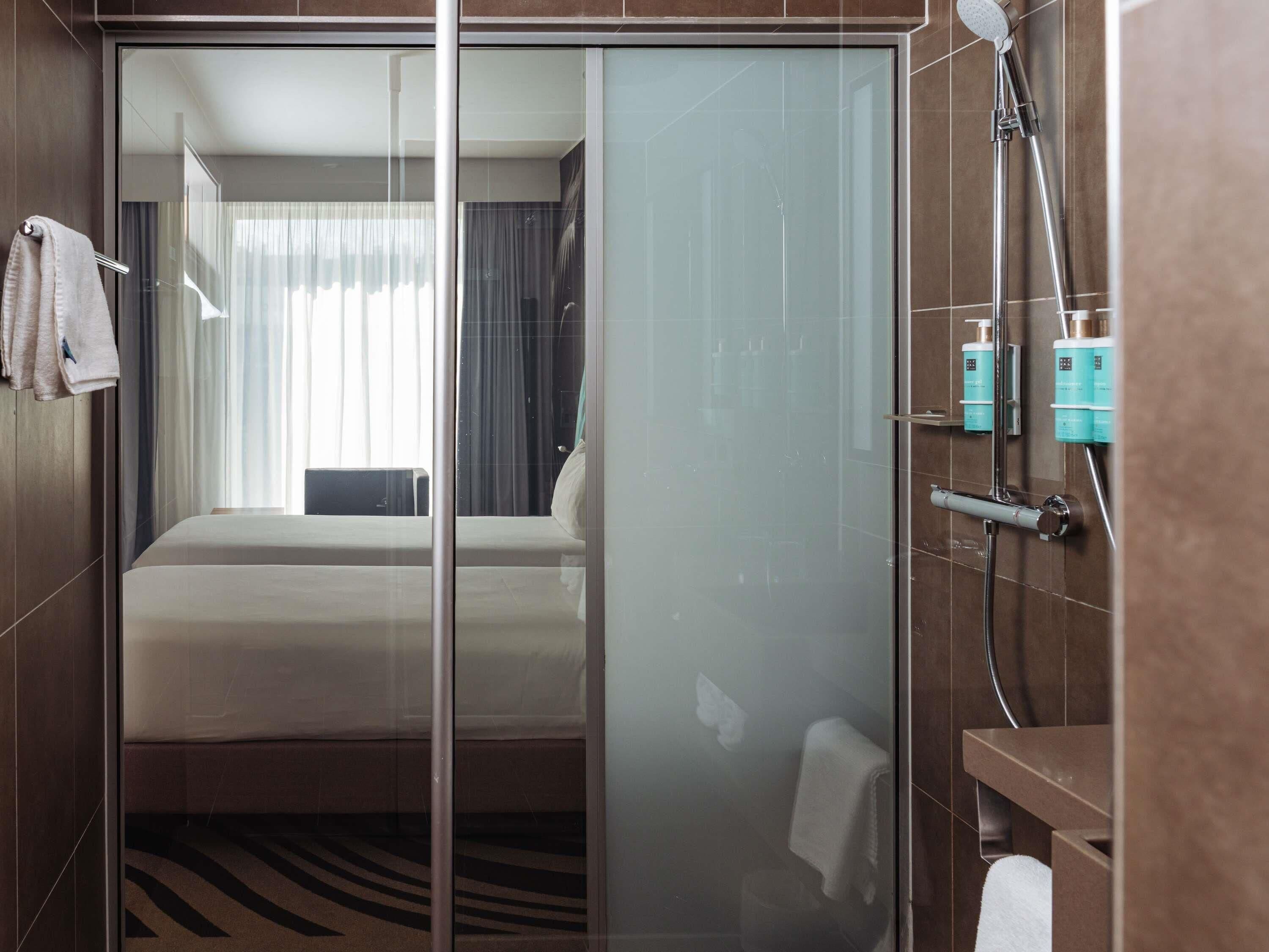 A hotel room with a glass partition separating the shower area from the bed.