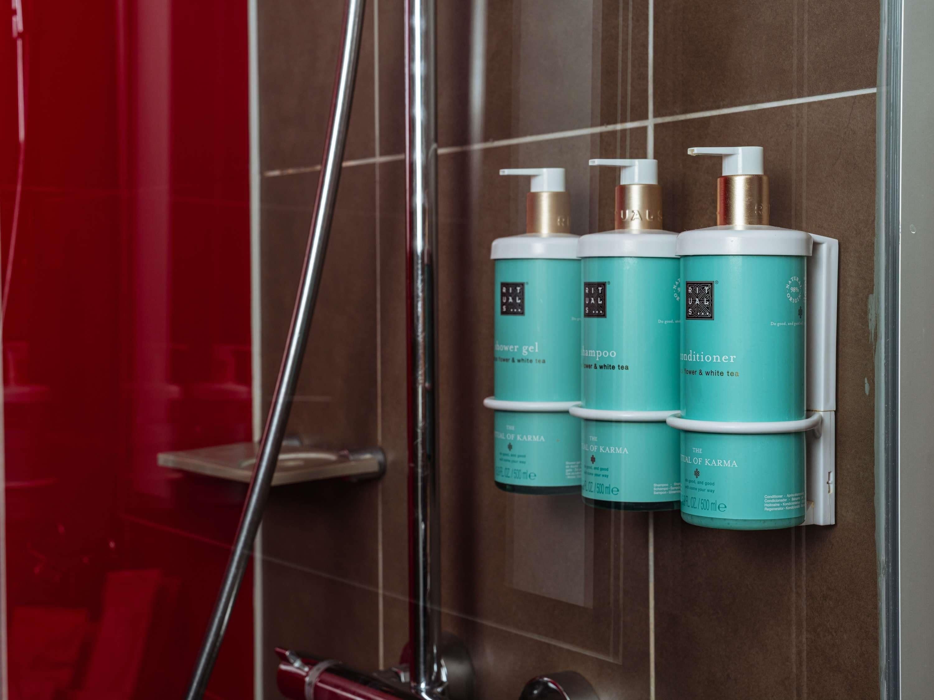 A bathroom with a shower and a wall-mounted rack holding four bottles of shower gel, shampoo, conditioner, and body wash.