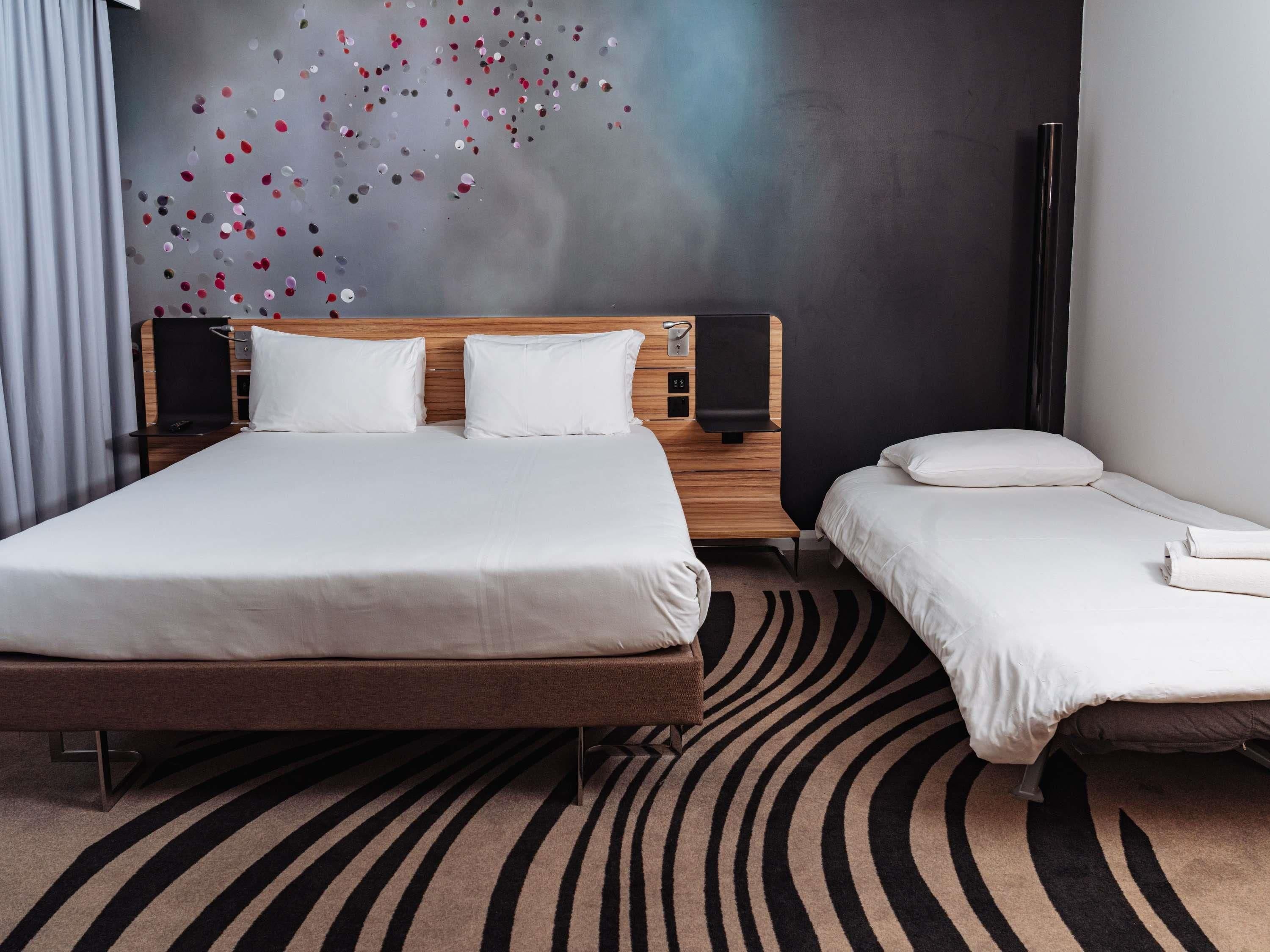 Een hotelkamer met een groot bed, een kleiner bed, een houten hoofdeinde, twee nachtkastjes, een wanddecoratie en een kamerkleed met dessin.