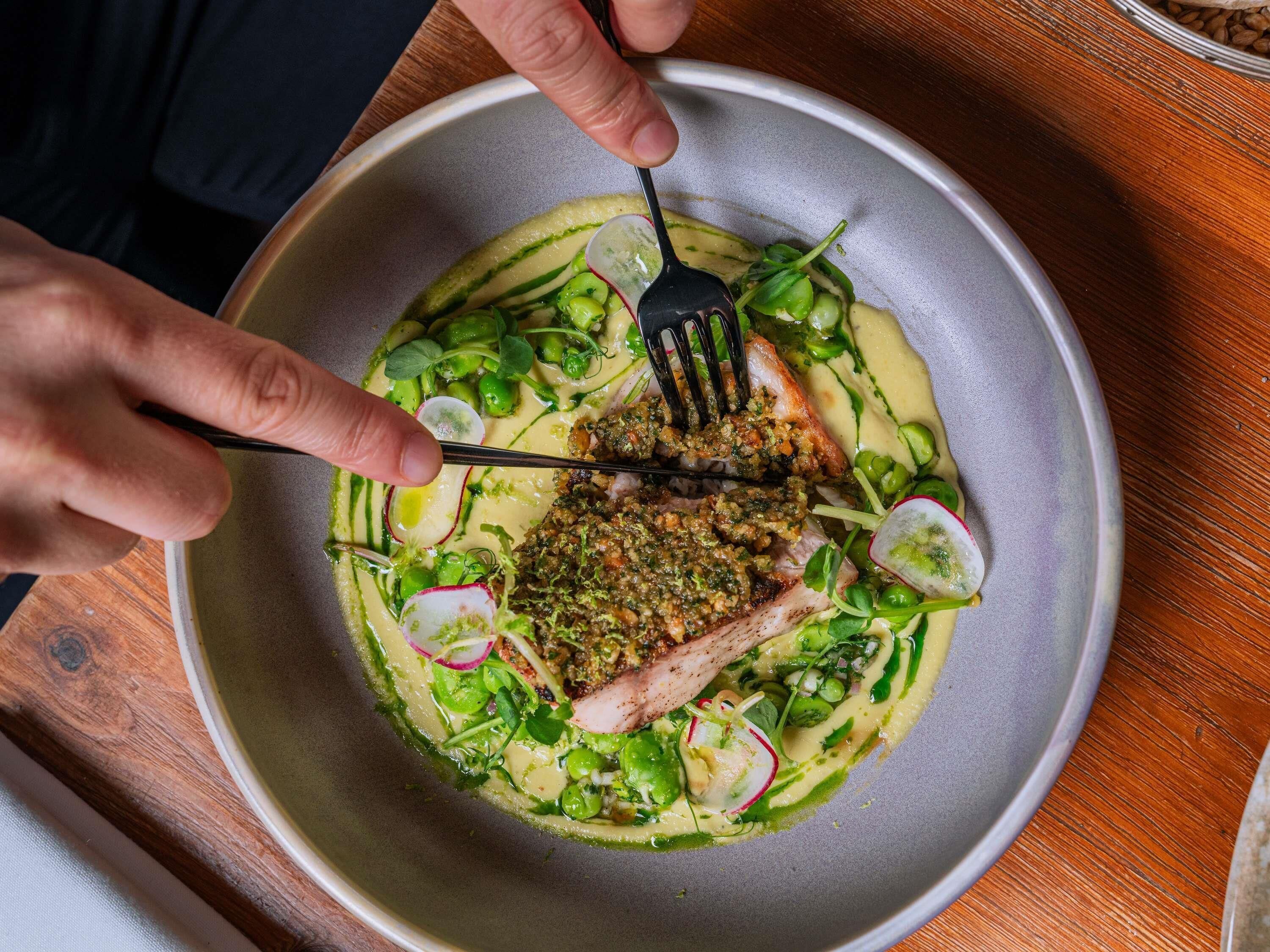 A person holding a fork over a plate with a piece of meat and green vegetables.