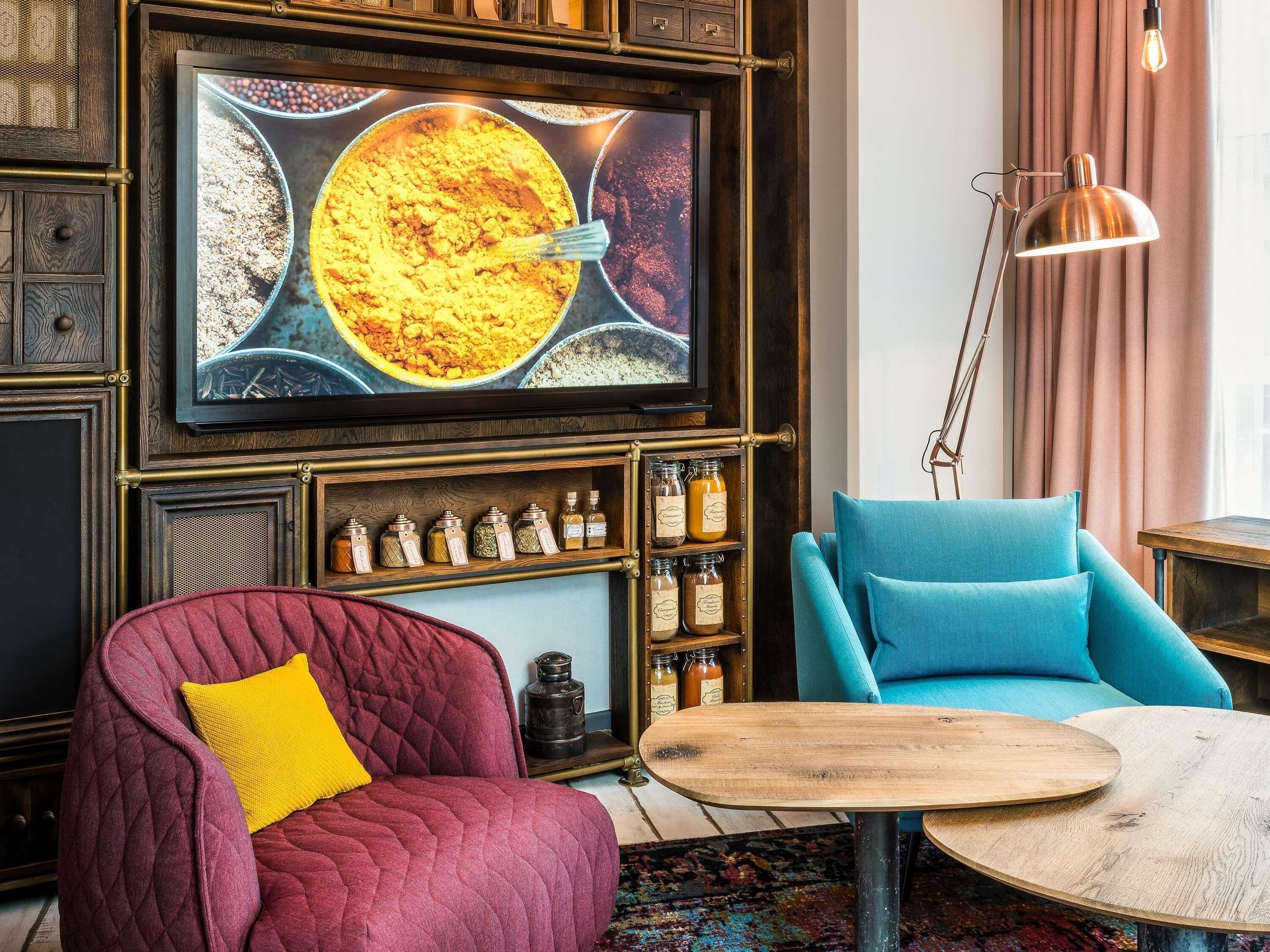 Una sala de estar acogedora con un sillón rojo, un sillón azul, una mesa de centro de madera, un televisor montado en la pared y un estante con frascos.