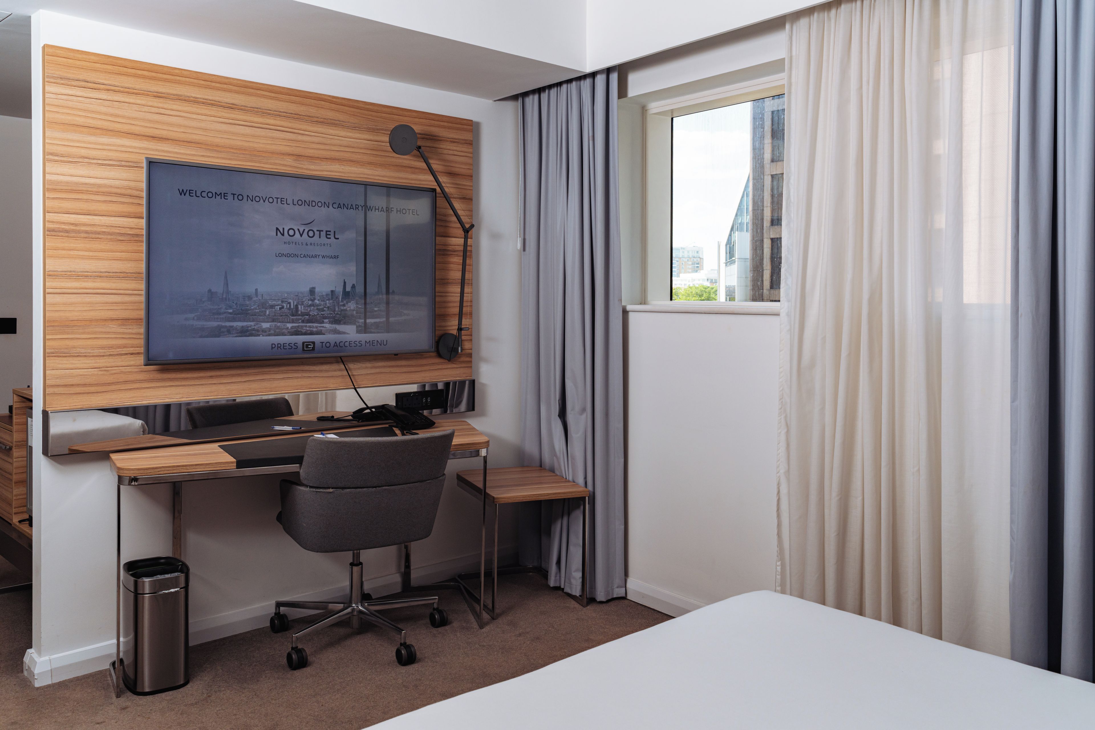 A hotel room with a wooden accent wall, a desk with a chair, a trash bin, and a window with curtains.