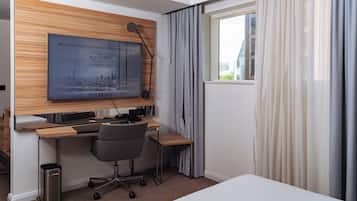 A hotel room with a wooden accent wall, a desk with a chair, a trash bin, and a window with curtains.