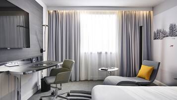 Une chambre d’hôtel moderne, équipée d’un lit, d’un bureau avec une chaise, d’une petite table et d’une grande fenêtre avec des rideaux.