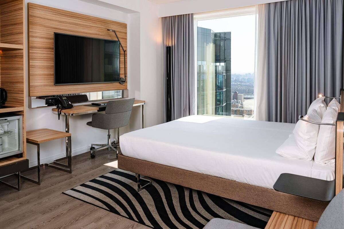 Une chambre d’hôtel moderne dotée d’un grand lit, d’un bureau avec une chaise, d’un téléviseur fixé au mur et offrant une vue sur la ville par la fenêtre.