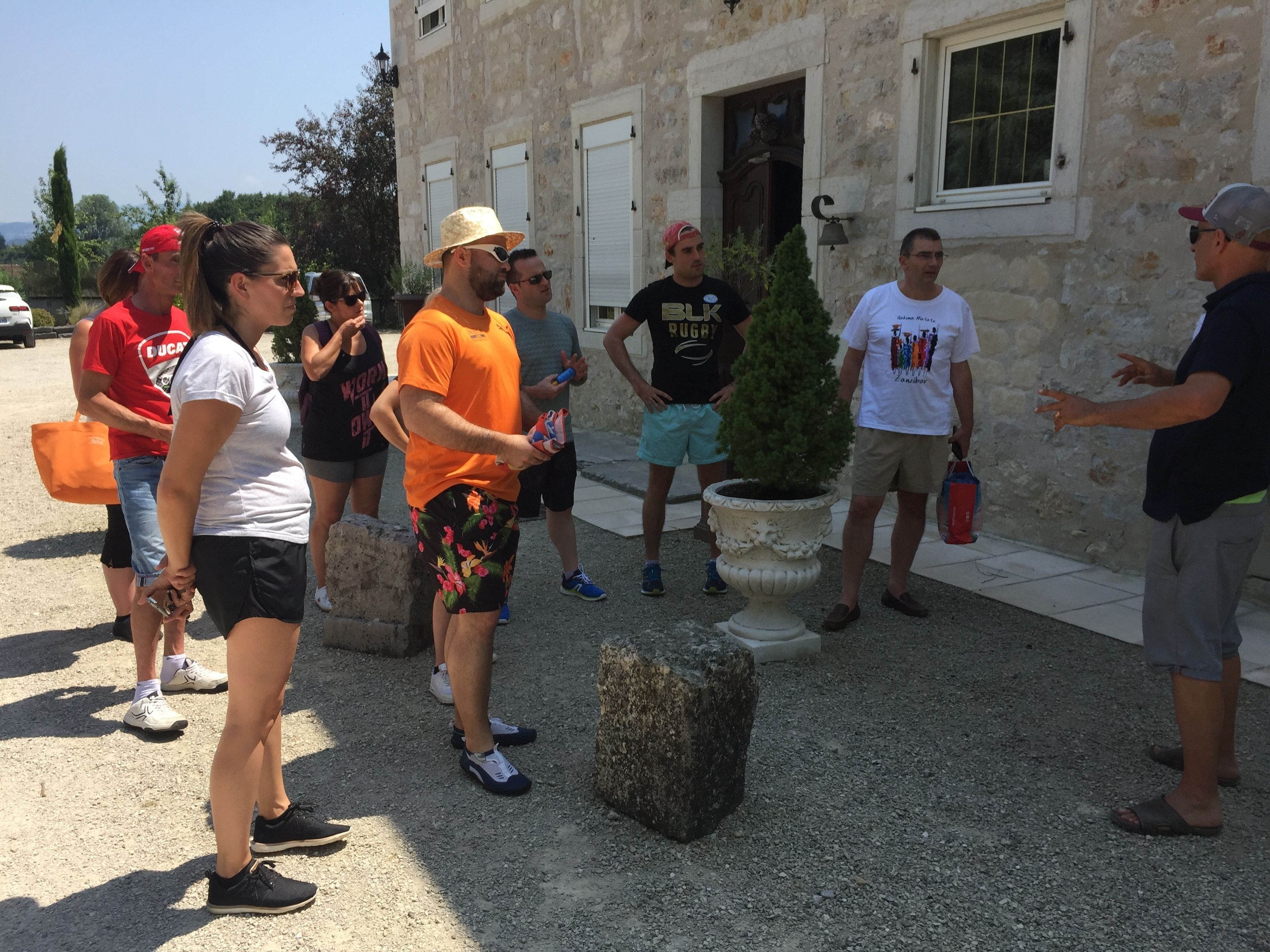 A group of people standing outdoors in front of a stone building with a large potted plant and a stone bench.