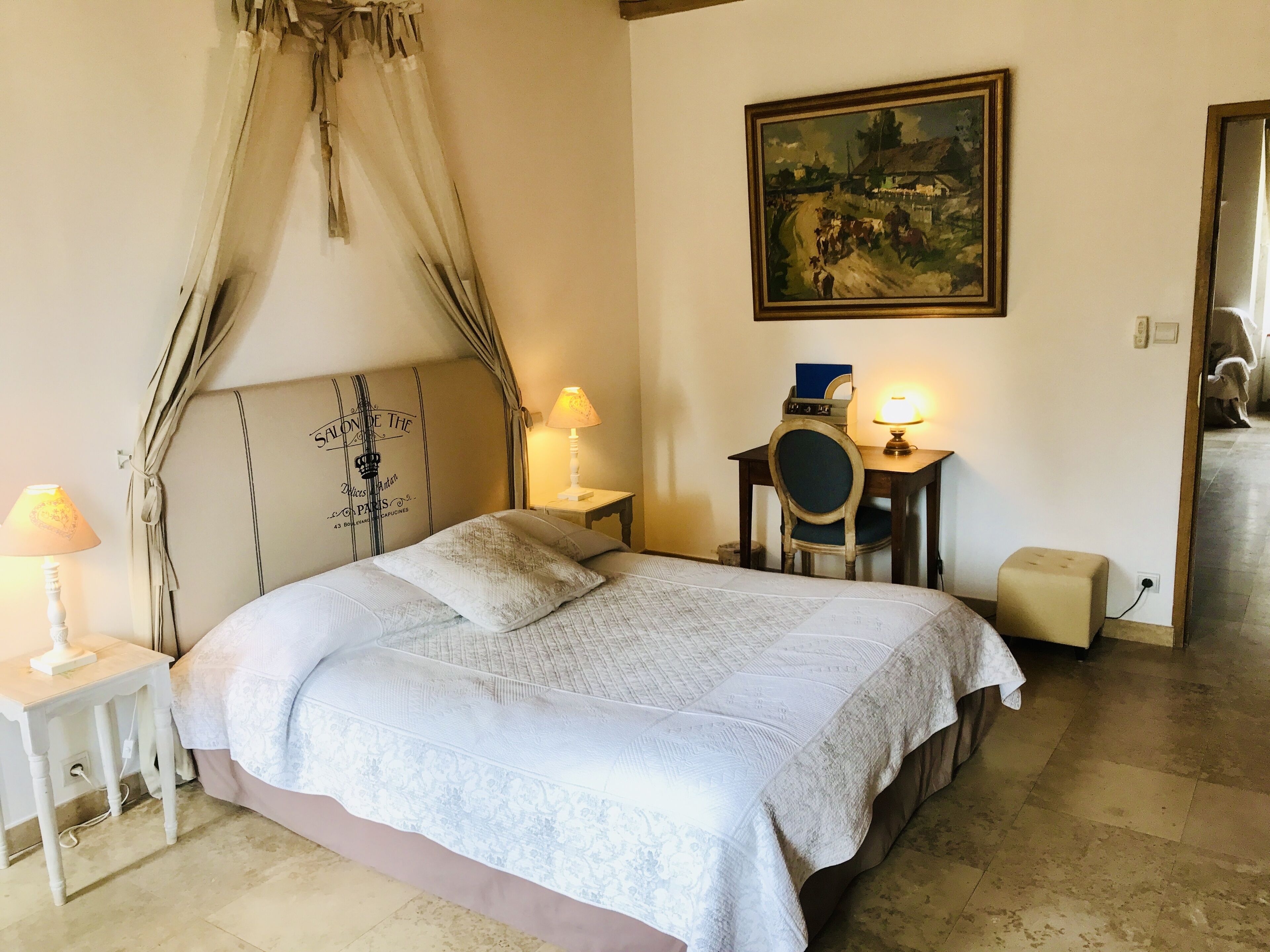 Una camera da letto ordinata con un letto, due lampade ai comodini, una scrivania con sedia, un piccolo tavolino e un quadro appeso al muro.