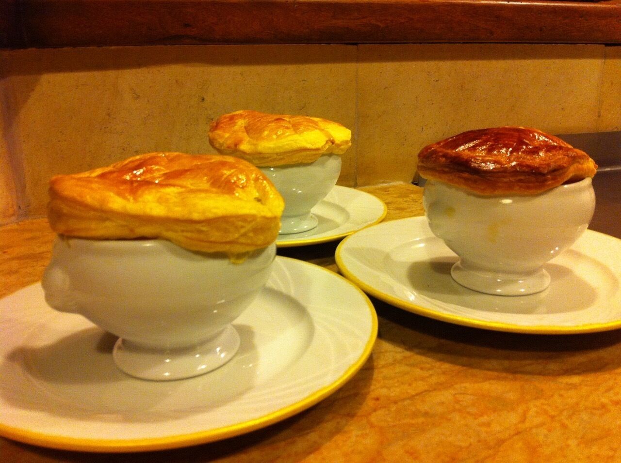 Tres tazones blancos de cerámica con tapas de pastelería doradas sobre platos blancos con detalles dorados, sobre una encimera de mármol.