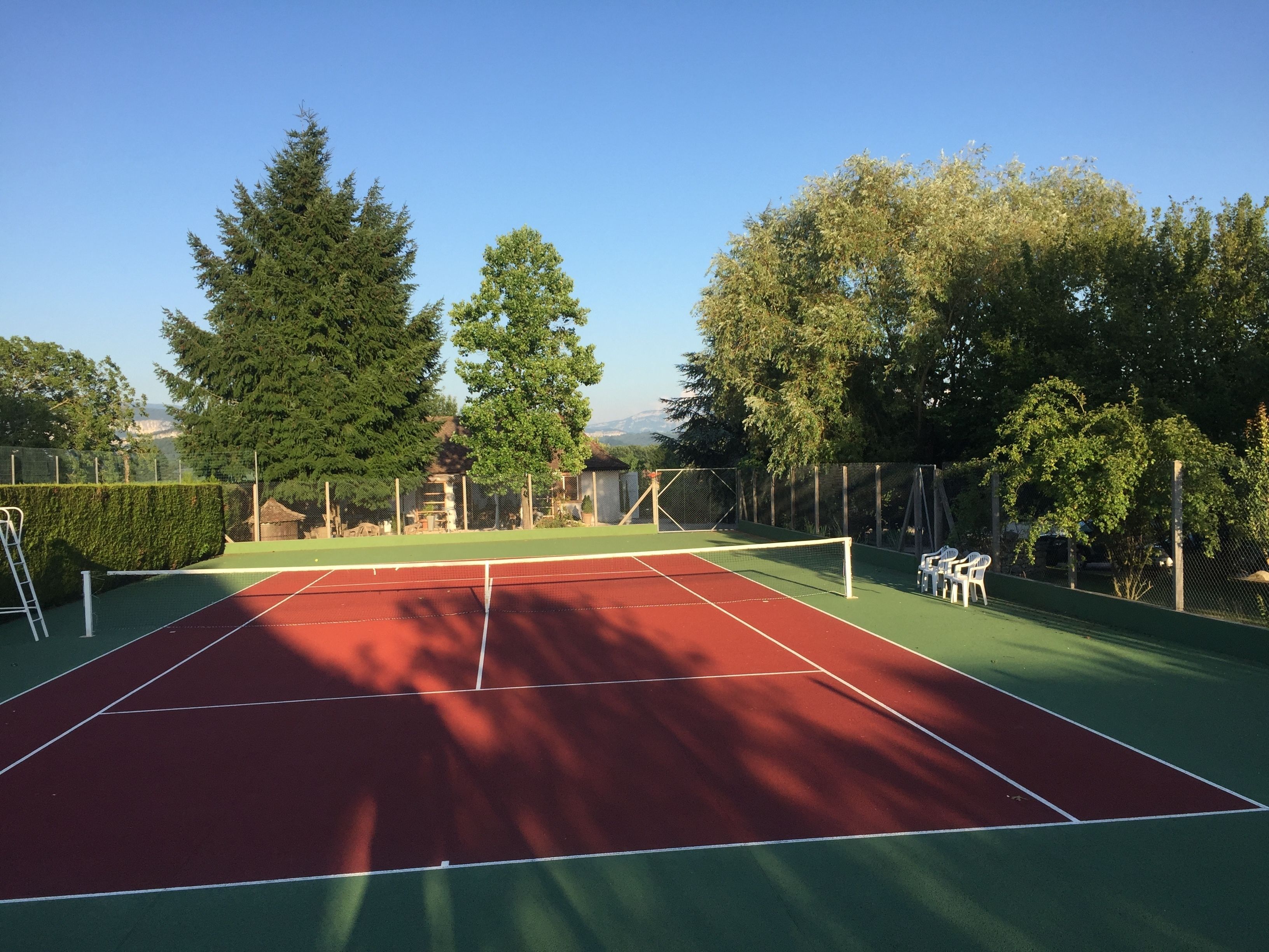 An outdoor tennis court with a net, surrounded by trees and a fence.
