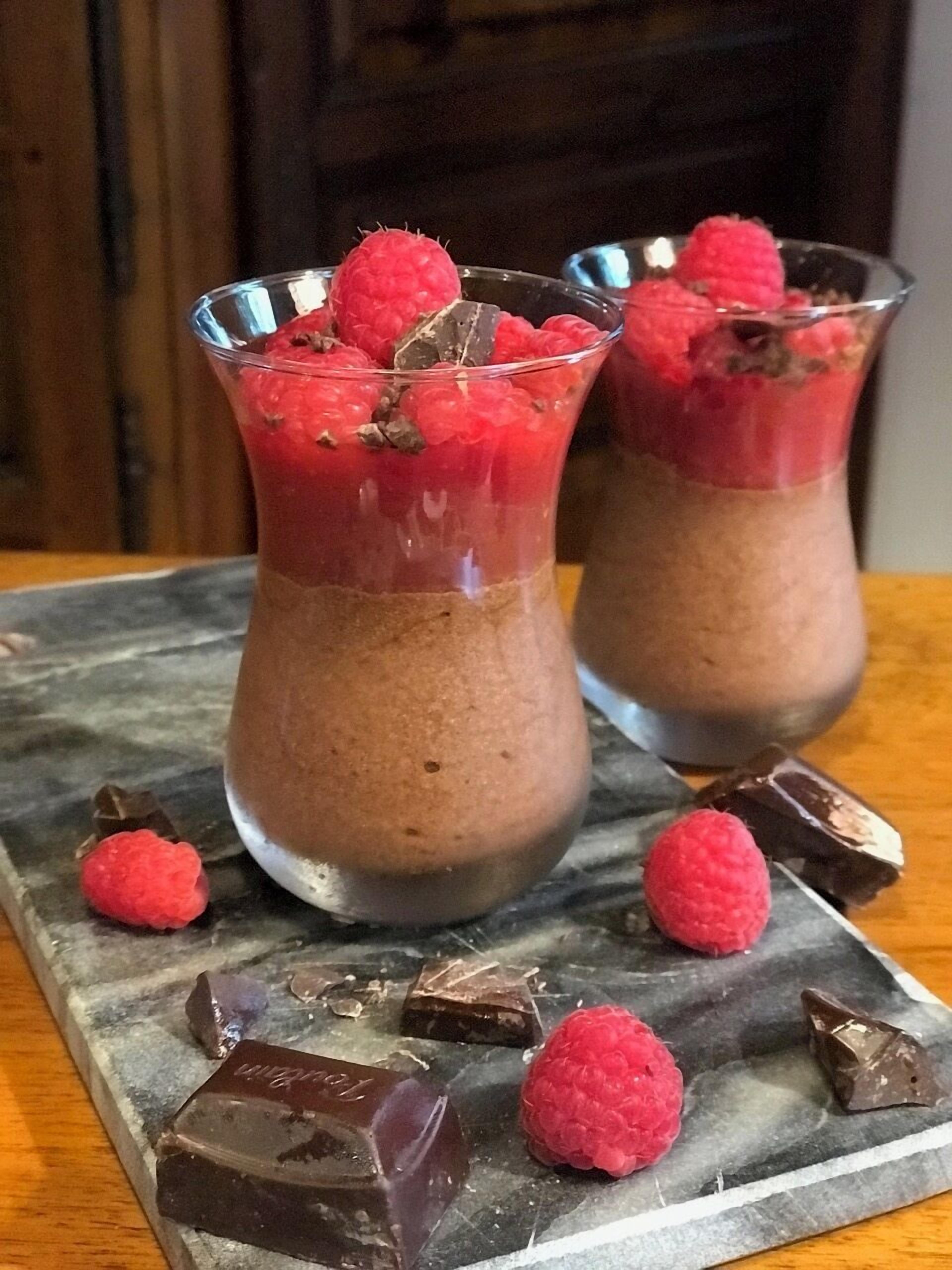 Dos vasos de mousse de chocolate cubiertos de frambuesas, sobre una superficie de mármol con trozos de chocolate esparcidos.