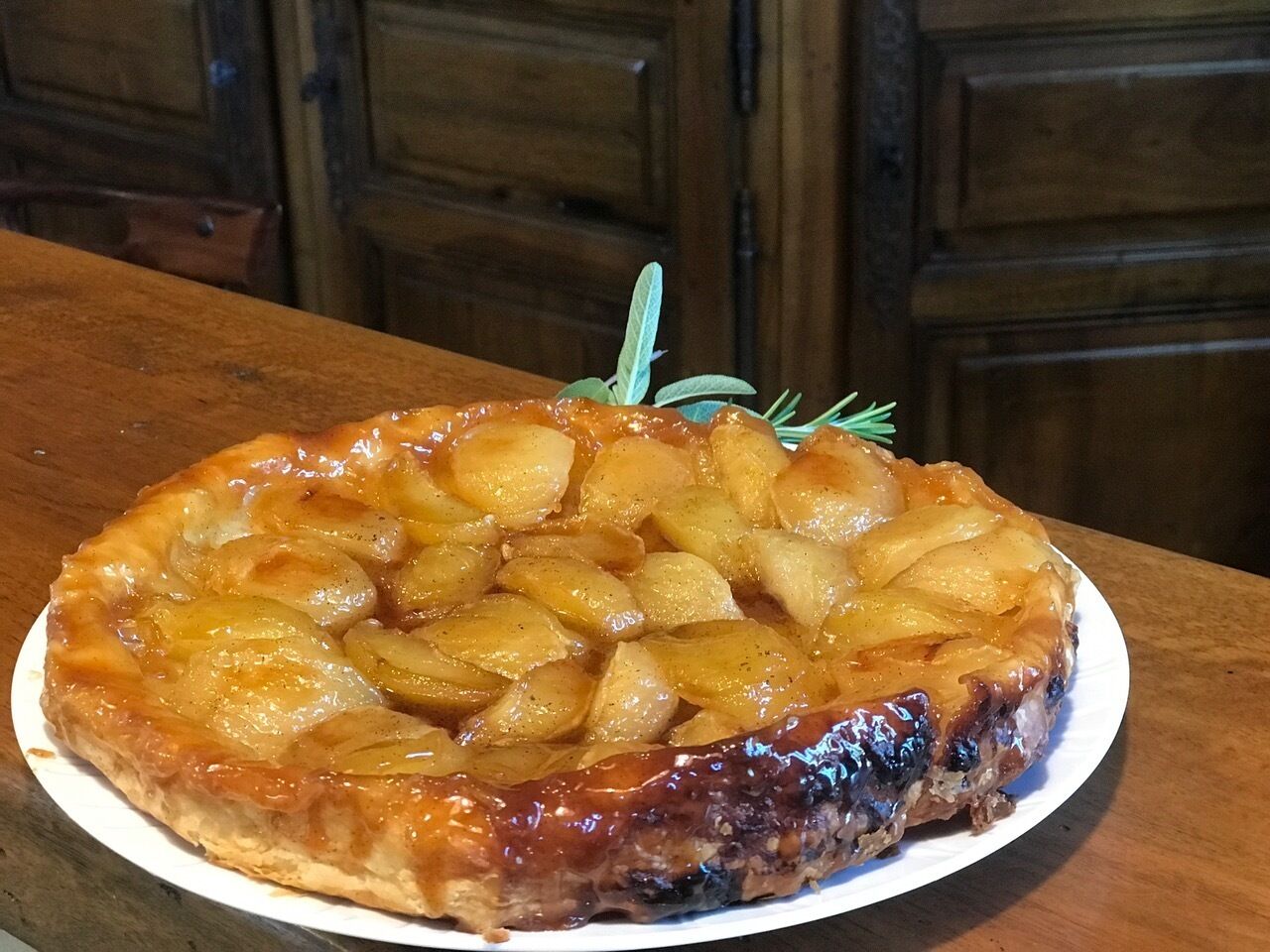 A round tart with a golden-brown crust, topped with sliced apples, and garnished with a sprig of rosemary.
