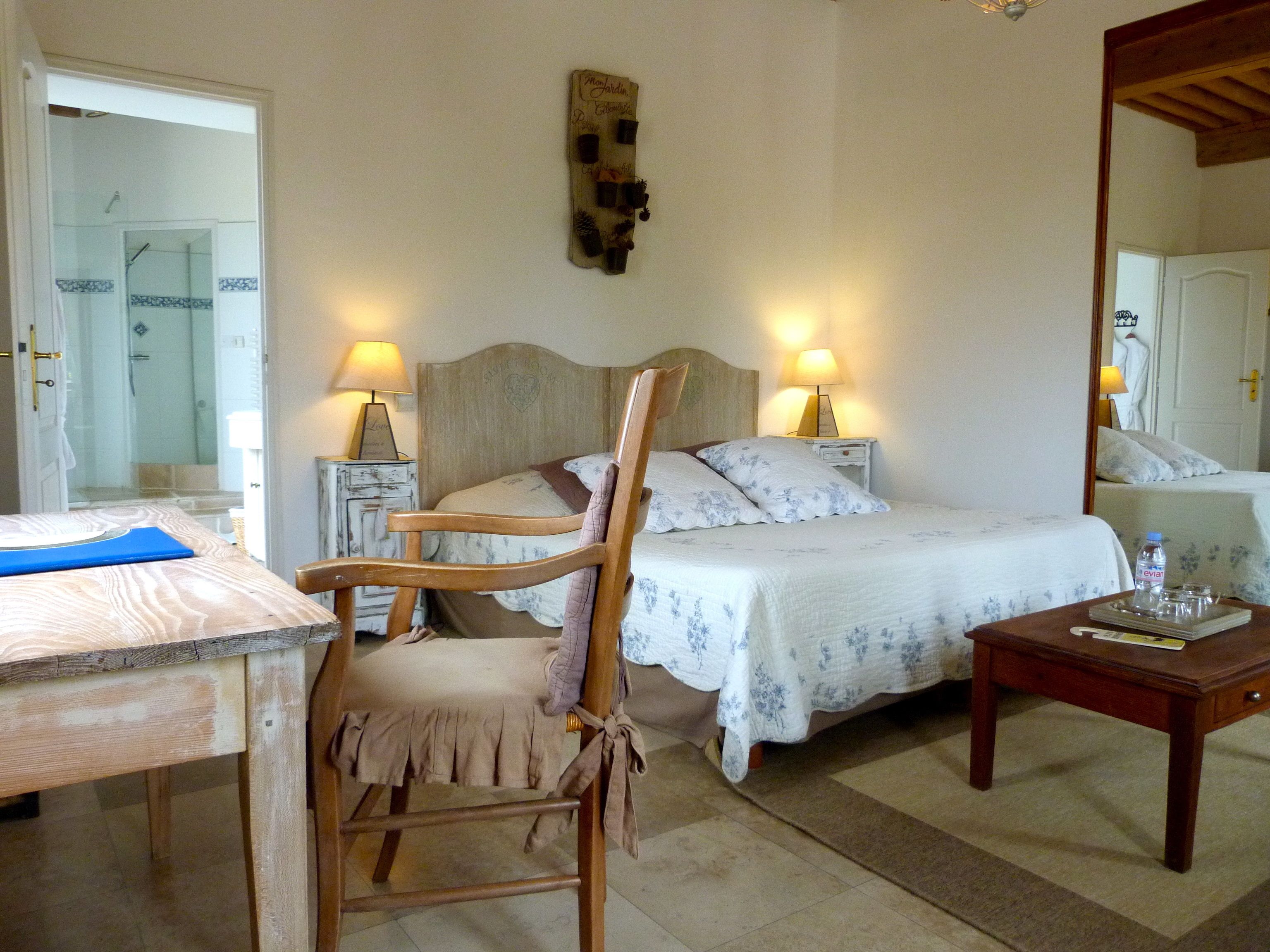 Une chambre à coucher avec un lit, deux tables de chevet avec des lampes, une chaise en bois, une petite table avec une bouteille et un livre, et un miroir au mur.