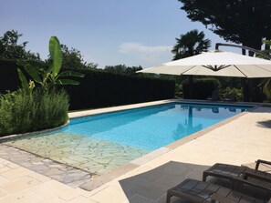 A swimming pool with tiled edges, surrounded by a patio area with chairs and an umbrella.