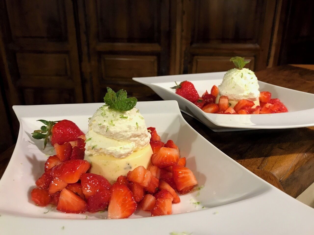 Two square white plates with a dessert consisting of a scoop of ice cream on top of a yellow base, surrounded by sliced strawberries.