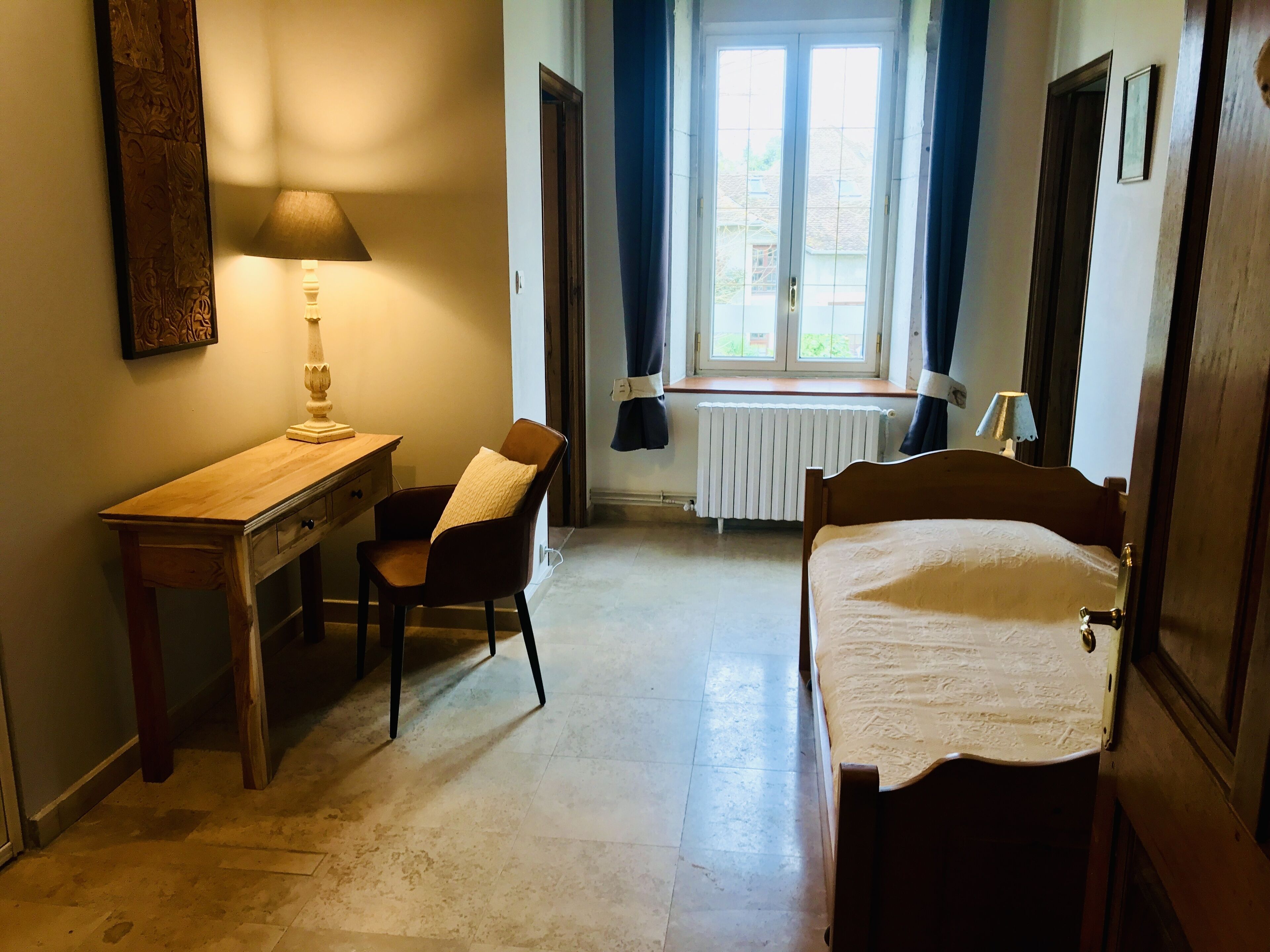 Una habitación pequeña y bien iluminada con escritorio de madera, una silla, una cama y un ventanal con cortinas.