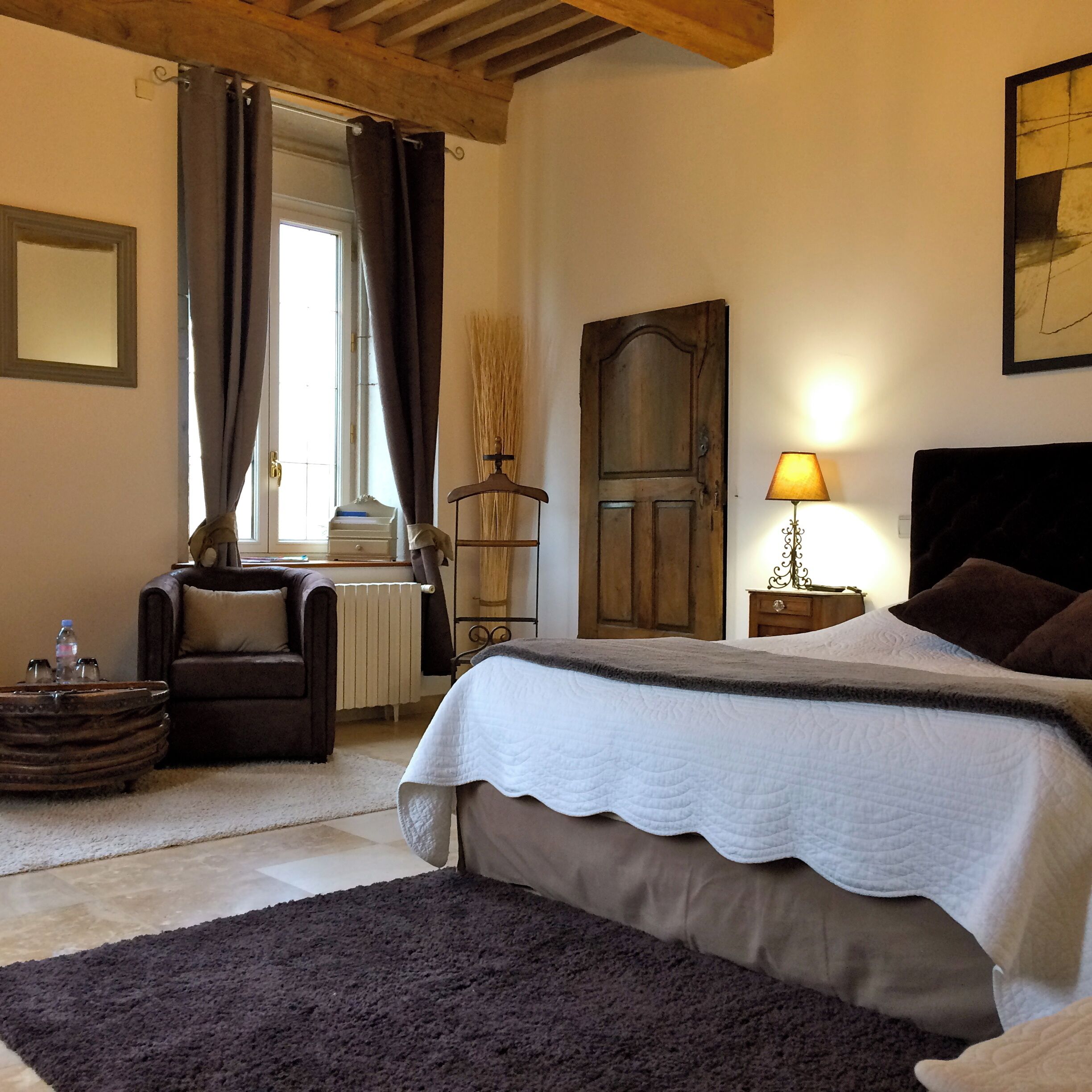 A bedroom with a large bed, a chair, a bedside table with a lamp, a radiator, and a window with curtains.