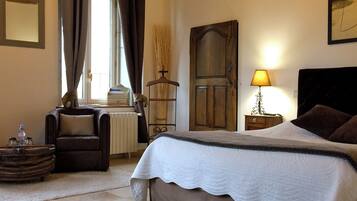 A bedroom with a large bed, a chair, a bedside table with a lamp, a radiator, and a window with curtains.