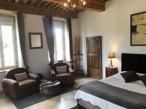 A bedroom with a bed, two leather armchairs, a wooden side table with a lamp, a mirror, and a door.