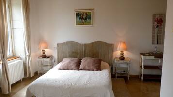 A bedroom with a bed, two bedside tables, a nightstand, a radiator, a window with curtains, a painting on the wall, and a desk with a lamp.